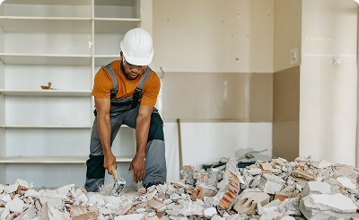 Construction worker clearing rubble indoors with a dust mask and helmet.