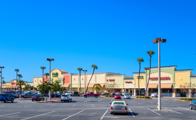 Shopping center parking lot under a clear blue sky with scattered cars.