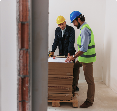 Two construction workers reviewing blueprints on a wooden pallet.